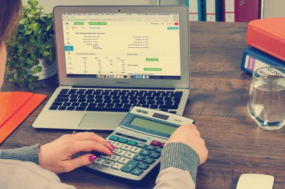 Hands using a calculator beside a laptop showing a spreadsheet
